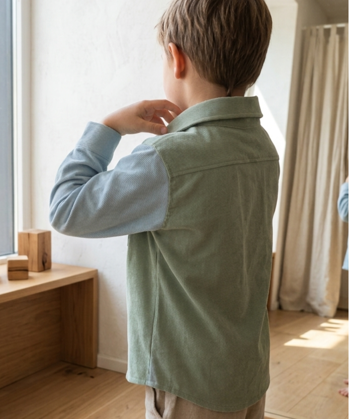 A child viewed from behind wearing a sage green corduroy overshirt with light blue long sleeves, standing in a sunlit room with wooden floors and a mirror.