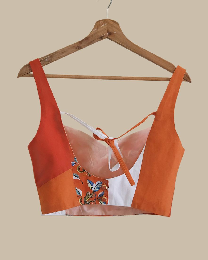 A women's crop top blouse with a color-blocked design in orange and white, featuring a monkey pattern on the back and a tie detail at the neckline, displayed on a wooden hanger against a solid beige background.
