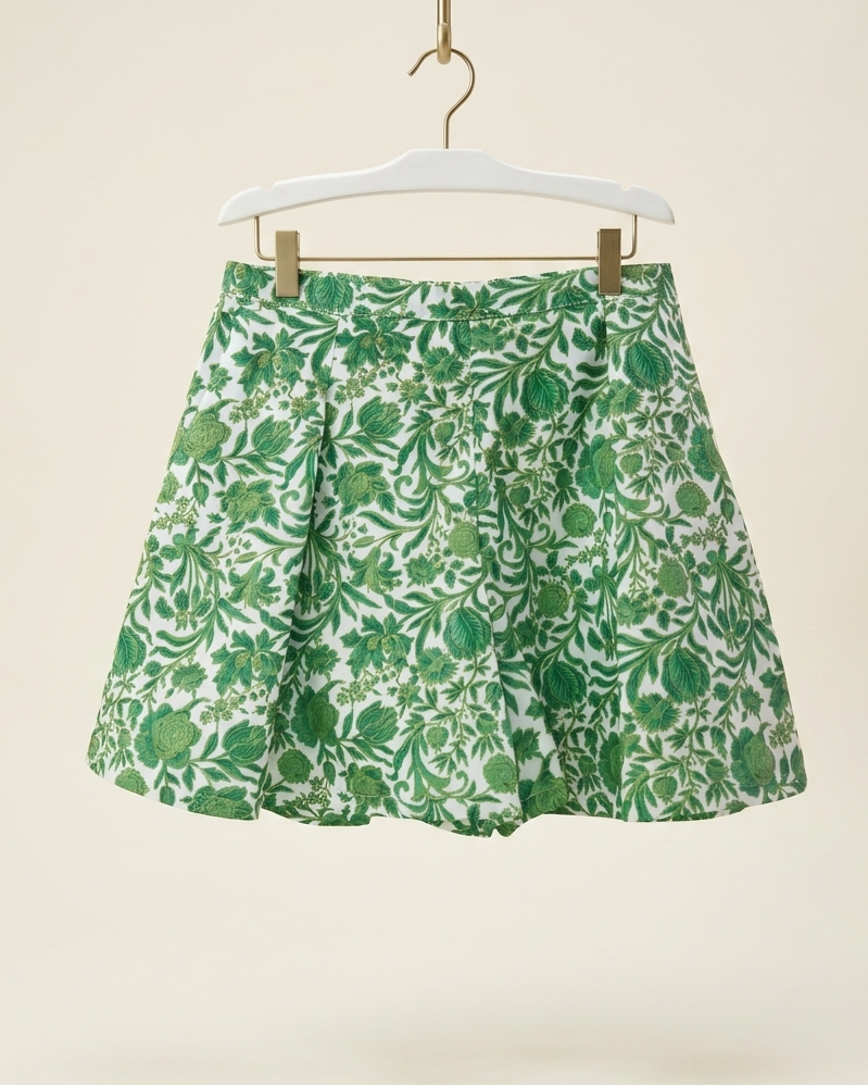 Green and Golden floral print pleated Short hanging on a white hanger with gold clips against a warm beige background.