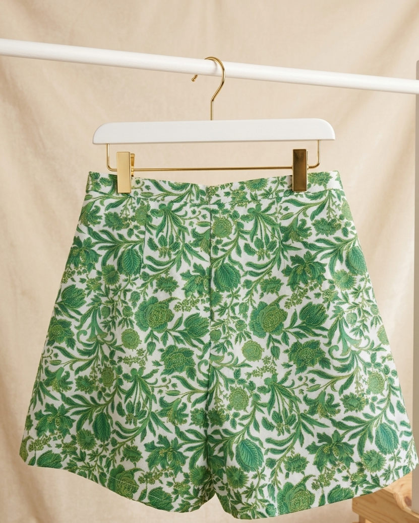 Green and white botanical print shorts hanging on a white hanger with gold clips against a beige fabric backdrop.