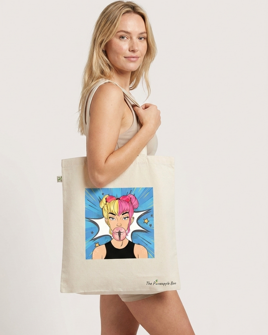 A woman modeling a cream canvas tote bag with a colorful pop-art graphic of a girl with pink and yellow hair.