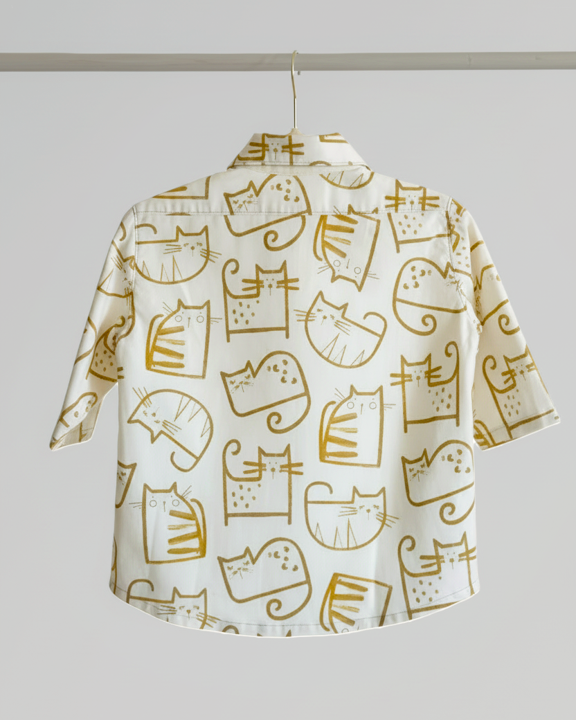 A short-sleeve button-up shirt with an all-over print of stylized cat faces and bodies in mustard yellow on a warm off-white background, hanging on a hanger against a light gray studio background.