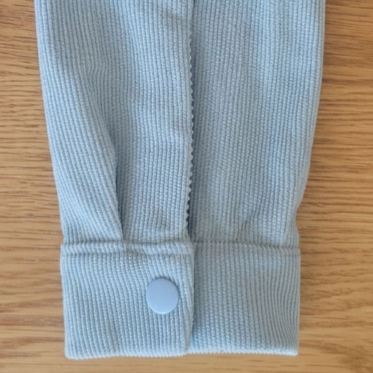 A close-up of a light blue shirt sleeve  with a single snap button closure, laid flat on a light brown wood surface.