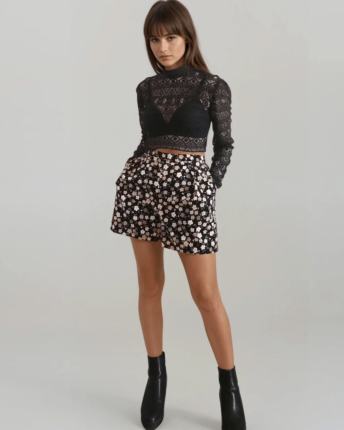 A model wearing a black long-sleeve lace mock-neck crop top and a black mini short with a white floral pattern, paired with black ankle boots, against a light gray studio background.